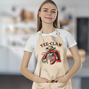 May include: A beige apron with a brown pocket and the text "YEE-CLAW" printed on the front. A cartoon lobster wearing a cowboy hat is holding a lasso.