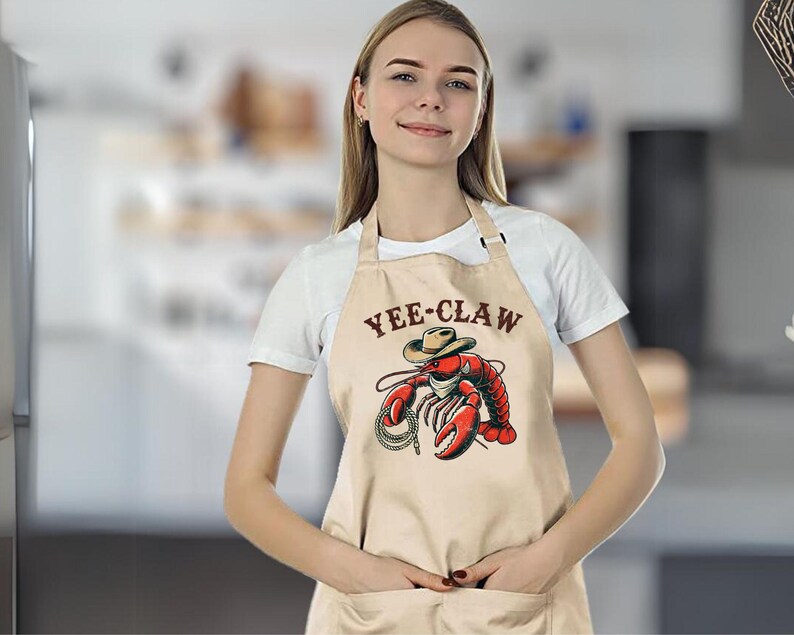 May include: A beige apron with a brown pocket and the text "YEE-CLAW" printed on the front. A cartoon lobster wearing a cowboy hat is holding a lasso.