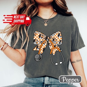 May include: Dark grey t-shirt featuring a large orange and white checkered bow with a stethoscope design. The bow has a pattern of a dog's face. The text "Pepper" is at the bottom.