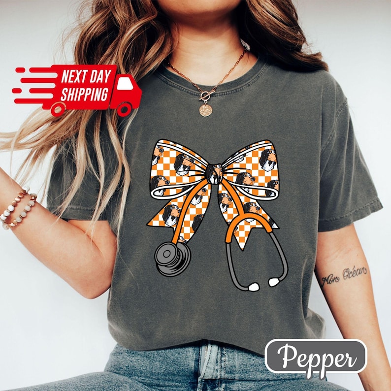 May include: Dark grey t-shirt featuring a large orange and white checkered bow with a stethoscope design. The bow has a pattern of a dog's face. The text "Pepper" is at the bottom.