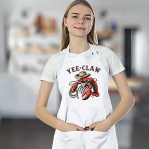 May include: White apron with a red lobster wearing a cowboy hat and the text "Yee-Claw".