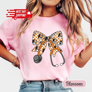 May include: Pink t-shirt featuring a bow design with a stethoscope. The bow has a checkered pattern with a dog's face. The stethoscope is orange and gray. The text "Next Day Shipping" is on the top left. The text "Blossom" is on the bottom right.