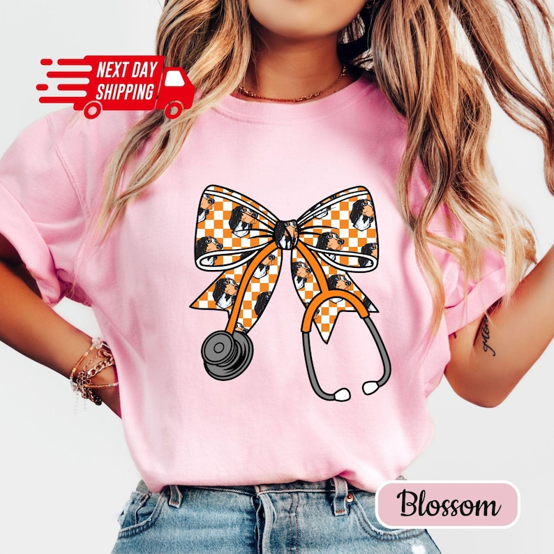 May include: Pink t-shirt featuring a bow design with a stethoscope. The bow has a checkered pattern with a dog's face. The stethoscope is orange and gray. The text "Next Day Shipping" is on the top left. The text "Blossom" is on the bottom right.