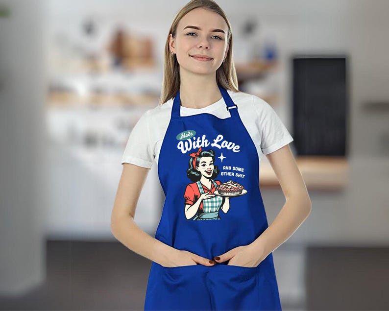 Puede incluir: Delantal azul real con un dise&ntilde;o retro de una mujer sosteniendo un pastel. El delantal tiene el texto "Made With Love and Some Other Shit". Cuenta con un bolsillo frontal y una correa para el cuello.