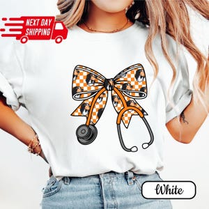 May include: White t-shirt featuring a bow and stethoscope design. The bow has a checkered pattern with dog faces and orange accents. The stethoscope is gray and orange. The shirt also has the text "Next Day Shipping" and the word "White".