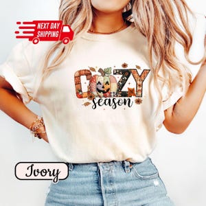 Op de afbeelding: Ivoor kleurig T-shirt met de tekst "COZY season" in een herfstthema ontwerp. Het ontwerp bevat een patchwork pompoen en herfstbladeren. Het shirt heeft korte mouwen en een ronde hals.