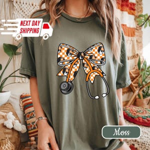 May include: Olive green t-shirt featuring a large bow design with a checkered pattern and dog faces. An orange stethoscope is overlaid on the bow. The shirt has the word "Moss" printed on the bottom right.