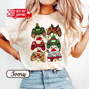 May include: Ivory-colored t-shirt featuring six Christmas-themed video game controller designs. Each controller is decorated with festive elements like a Christmas tree, reindeer antlers, Santa hats, and a snowman. The shirt also has the text "Next Day Shipping".