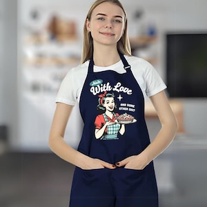 Puede incluir: Delantal azul marino con una ilustraci&oacute;n retro de una mujer sosteniendo un pastel. El delantal tiene el texto "Made With Love and some other shit". Cuenta con un peto, bolsillos y una correa para el cuello. El delantal es usado por una persona.