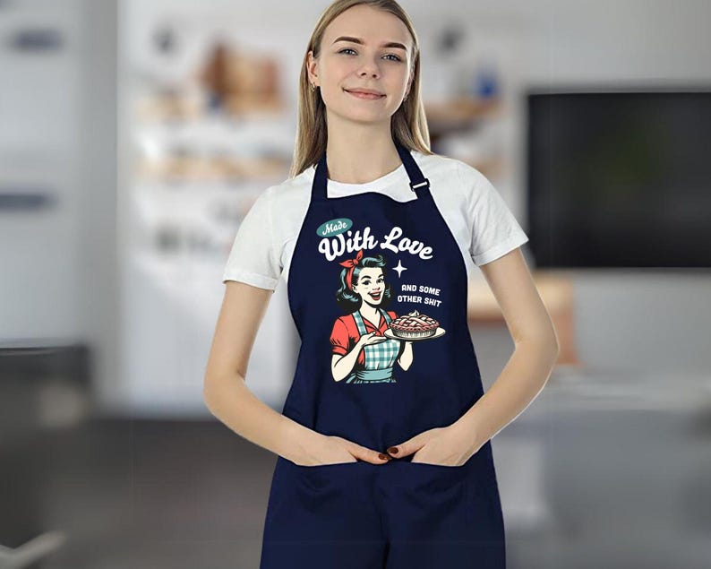 Puede incluir: Delantal azul marino con una ilustraci&oacute;n retro de una mujer sosteniendo un pastel. El delantal tiene el texto "Made With Love and some other shit". Cuenta con un peto, bolsillos y una correa para el cuello. El delantal es usado por una persona.
