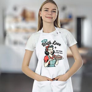 Puede incluir: Delantal blanco con una ilustraci&oacute;n retro de una mujer sosteniendo un pastel. El delantal tiene el texto "Made With Love and some other shit" en un estilo vintage. El delantal tiene un bolsillo.
