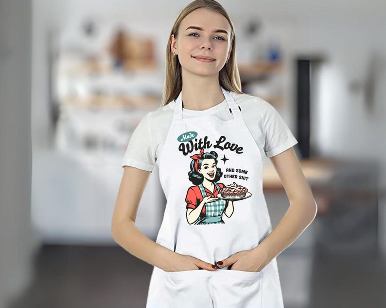 Puede incluir: Delantal blanco con una ilustraci&oacute;n retro de una mujer sosteniendo un pastel. El delantal tiene el texto "Made With Love and some other shit" en un estilo vintage. El delantal tiene un bolsillo.