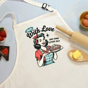 Puede incluir: Delantal blanco con una ilustraci&oacute;n retro de una mujer sosteniendo un pastel, con el texto "Made With Love and some other shit." El delantal tiene una correa blanca y una hebilla negra. Tambi&eacute;n se ven fresas, chocolate, huevos, un rodillo y mantequilla.