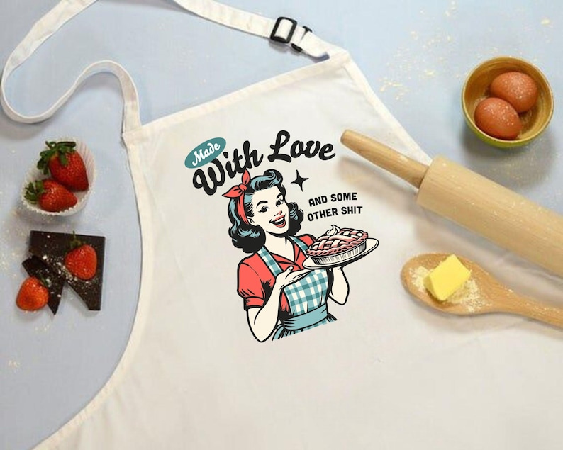 Puede incluir: Delantal blanco con una ilustraci&oacute;n retro de una mujer sosteniendo un pastel, con el texto "Made With Love and some other shit." El delantal tiene una correa blanca y una hebilla negra. Tambi&eacute;n se ven fresas, chocolate, huevos, un rodillo y mantequilla.