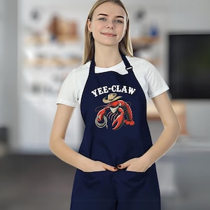 May include: A navy blue apron with a white graphic of a lobster wearing a cowboy hat and the text "YEE-CLAW".