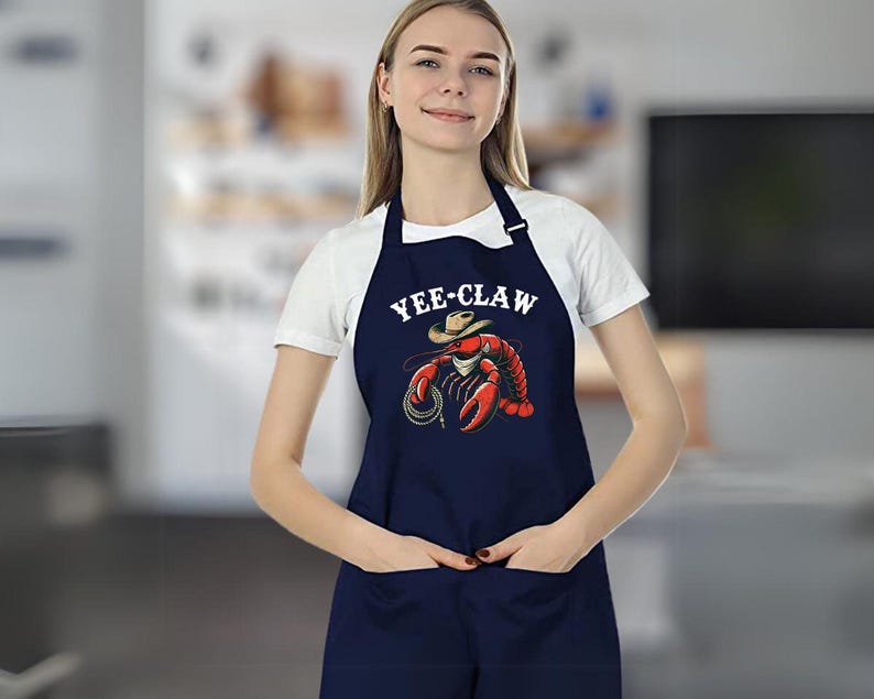 May include: A navy blue apron with a white graphic of a lobster wearing a cowboy hat and the text "YEE-CLAW".