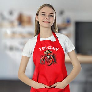 May include: Red kitchen apron with a white graphic of a lobster wearing a cowboy hat and the text "Yee-Claw".