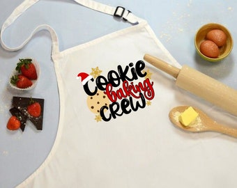 Może przedstawiać: Biały fartuch z napisem "Cookie Baking Crew" w czarnych i czerwonych literach. Ciasteczko piernikowe z czerwonym kapeluszem Świętego Mikołaja znajduje się w środku tekstu. Wokół tekstu znajdują się złote płatki śniegu.