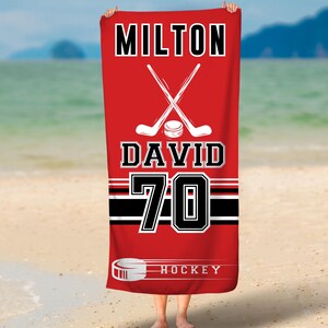 May include: Red beach towel with a hockey stick and puck design. The towel has the text "MILTON DAVID 70 HOCKEY" printed on it.