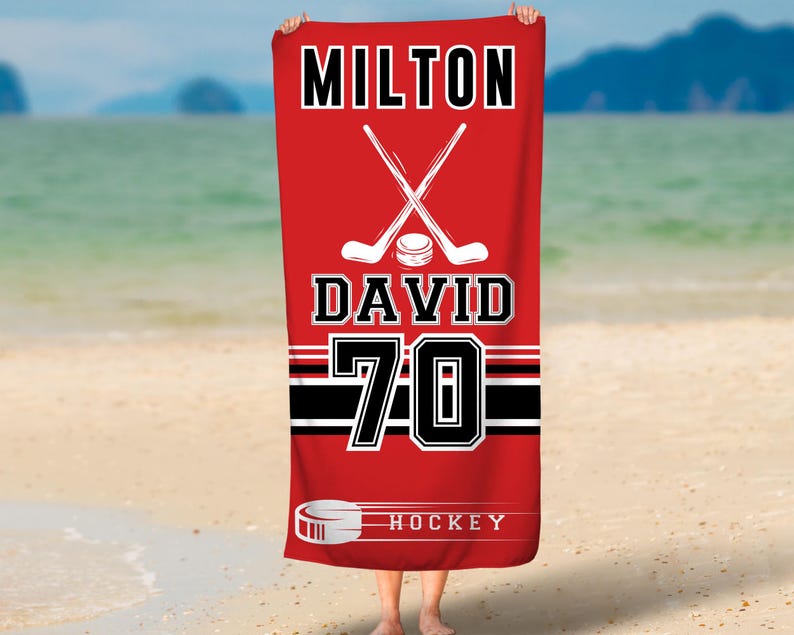 May include: Red beach towel with a hockey stick and puck design. The towel has the text "MILTON DAVID 70 HOCKEY" printed on it.
