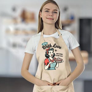 Puede incluir: Delantal beige con una ilustraci&oacute;n retro de una mujer sosteniendo un pastel. El delantal tiene el texto "Made With Love and Some Other Shit". La mujer de la ilustraci&oacute;n lleva un pa&ntilde;uelo rojo y una blusa roja.