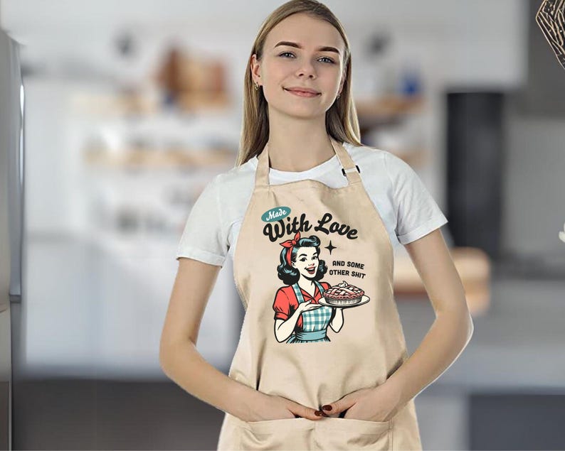 Puede incluir: Delantal beige con una ilustraci&oacute;n retro de una mujer sosteniendo un pastel. El delantal tiene el texto "Made With Love and Some Other Shit". La mujer de la ilustraci&oacute;n lleva un pa&ntilde;uelo rojo y una blusa roja.