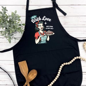 Puede incluir: Delantal negro con un bolsillo frontal grande y una correa ajustable para el cuello. El delantal presenta una ilustraci&oacute;n retro de una mujer sosteniendo un pastel, con el texto "Made With Love AND SOME OTHER SHIT". Cucharas de madera y una planta en el fondo.