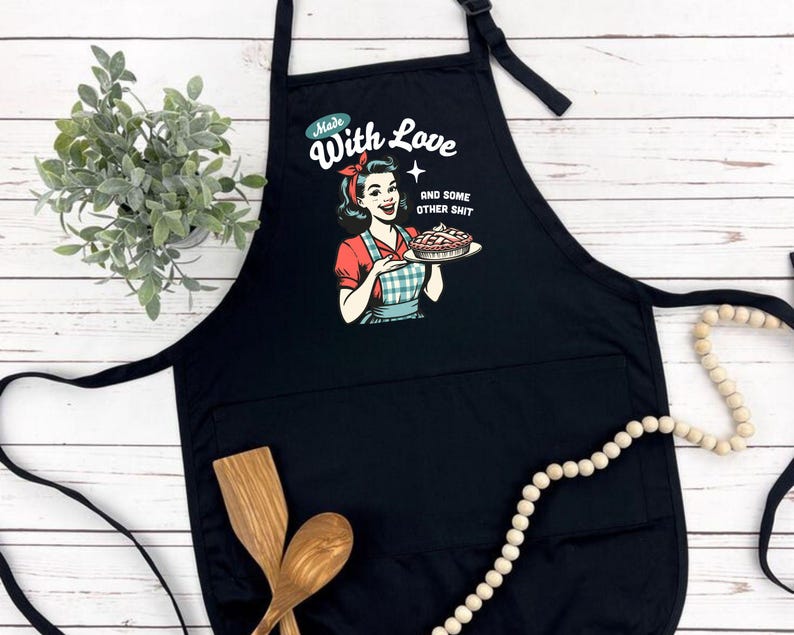 Puede incluir: Delantal negro con un bolsillo frontal grande y una correa ajustable para el cuello. El delantal presenta una ilustraci&oacute;n retro de una mujer sosteniendo un pastel, con el texto "Made With Love AND SOME OTHER SHIT". Cucharas de madera y una planta en el fondo.