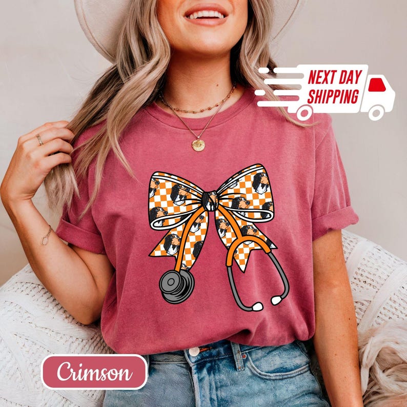 May include: Crimson-colored t-shirt featuring a bow design with a checkered pattern and a stethoscope. The bow incorporates a portrait, and the stethoscope has orange accents. The shirt also has the text "Crimson".
