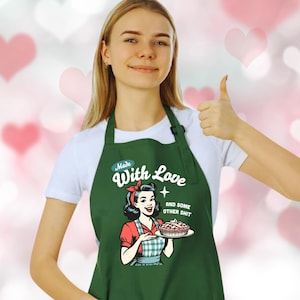 Puede incluir: Un delantal verde oscuro con el texto "Made With Love and some other shit". El delantal presenta una ilustraci&oacute;n retro de una mujer sosteniendo un pastel. La mujer lleva una blusa roja y un delantal a cuadros azul y blanco.