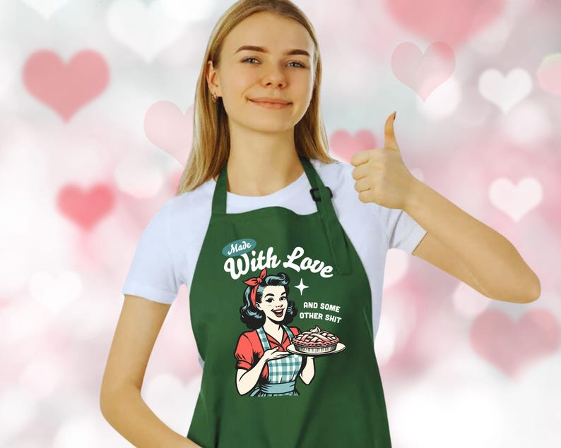 Puede incluir: Un delantal verde oscuro con el texto "Made With Love and some other shit". El delantal presenta una ilustraci&oacute;n retro de una mujer sosteniendo un pastel. La mujer lleva una blusa roja y un delantal a cuadros azul y blanco.
