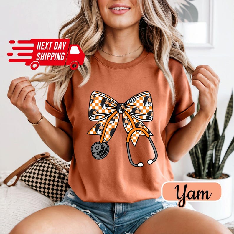 May include: A burnt orange t-shirt featuring a bow and stethoscope design. The bow has a checkered pattern with a dog's face, and the stethoscope is orange and gray. The shirt also has the text "Yam".