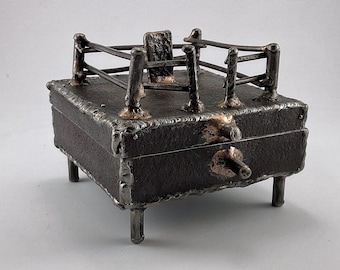 May include: A handcrafted metal box with a unique design. The dark metal box has a square shape, supported by four legs. The top features a railing-like structure. The box has a latch and is made with visible welding marks.