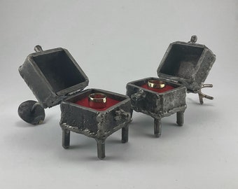 May include: Two rustic metal ring boxes with red velvet interiors. Each box has a gold ring inside and sits on four legs.