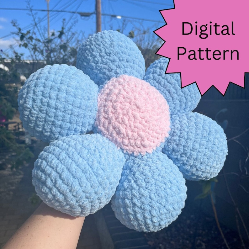 May include: A crocheted flower pillow in light blue and pink. The pillow is shaped like a flower with six petals. The center of the flower is pink and the petals are light blue. The pillow is a digital pattern.