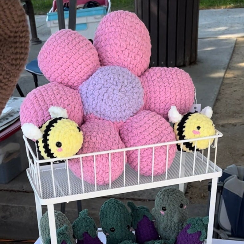May include: A crocheted flower-shaped pillow with a purple center and pink petals. Two small crocheted bee toys are on the shelf with the pillow.