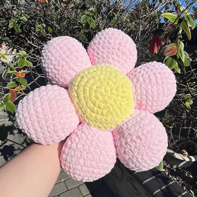 May include: A pink and yellow crocheted flower pillow. The pillow is shaped like a flower with six petals and a yellow center.