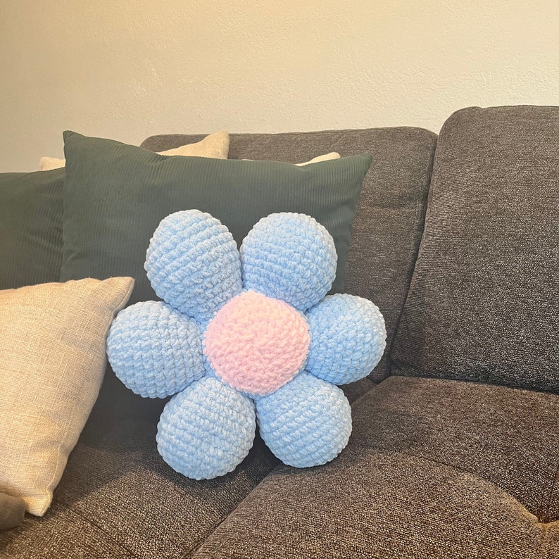May include: A light blue crocheted flower-shaped pillow with a pink center. The pillow is on a gray couch with green and brown throw pillows.