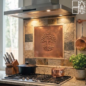 May include: A kitchen backsplash featuring a square copper panel embossed with a tree of life design, surrounded by multi-coloured natural stone tiles. A stainless steel range hood is mounted above a gas stovetop with a black pot and copper pot.