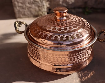 May include: A copper serving dish with a lid, featuring intricate engraved designs. The dish has two ornate handles and a polished finish.