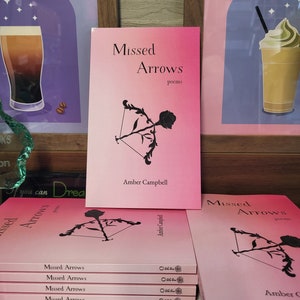 A stack of the book, "Missed Arrows: Poems," on a desk. The cover is a pink ombre with black details and features a bow shooting a rose as an arrow.