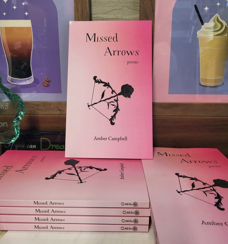 A stack of the book, "Missed Arrows: Poems," on a desk. The cover is a pink ombre with black details and features a bow shooting a rose as an arrow.