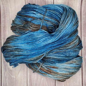 May include: A skein of blue yarn with brown and gray accents. The yarn is tightly wound and tied with a thin string. The yarn is on a light brown wooden surface.