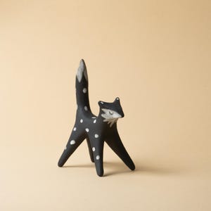 May include: A black fox ornament decorated with white spots. The fox has a tall tail and a pointed face. The fox's face and tail tip are painted white. The ornament is standing on a plain, light-coloured surface.