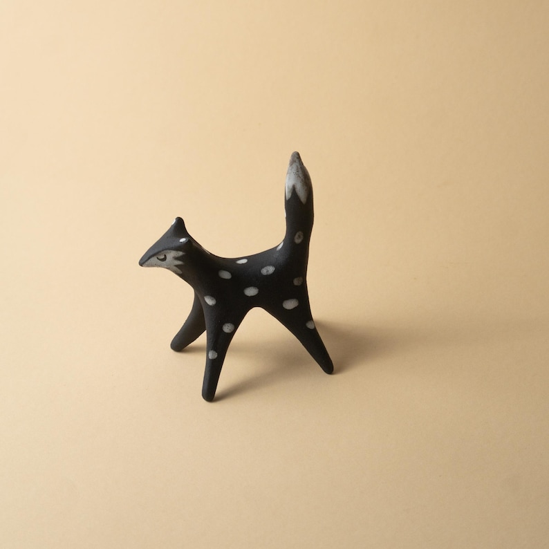 May include: A black ceramic fox figurine with white polka dots. The fox has a pointed snout, upright ears, and a tail with a white tip. The figurine is standing on four legs, with a minimalist design.