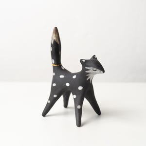May include: A black fox ornament with white spots and a gold collar. The fox has a pointed tail and ears, and a white face. The ornament is standing on four legs.
