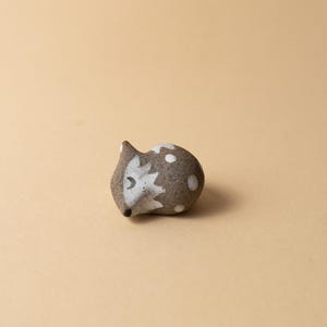 Op de afbeelding: Een klein keramisch egelfiguur. De egel is voornamelijk grijs met witte stippen en een wit gezicht. Het gezicht heeft een maanvormig oog en een zwarte neus. Een decoratief item.