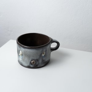 Op de afbeelding: Een brede, lage keramische mok met een donkerbruine binnenkant en een gespikkelde, getextureerde buitenglazuur in grijze en blauwe tinten. Het oppervlak heeft kleine, verhoogde, onregelmatige uitsteeksels en een enkel handvat aan de rechterkant.