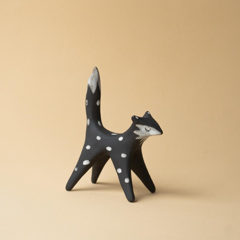 May include: A black ceramic fox figurine with white polka dots. The fox has a white-tipped tail and a white face with closed eyes. The figurine is standing on four legs and has a minimalist design.