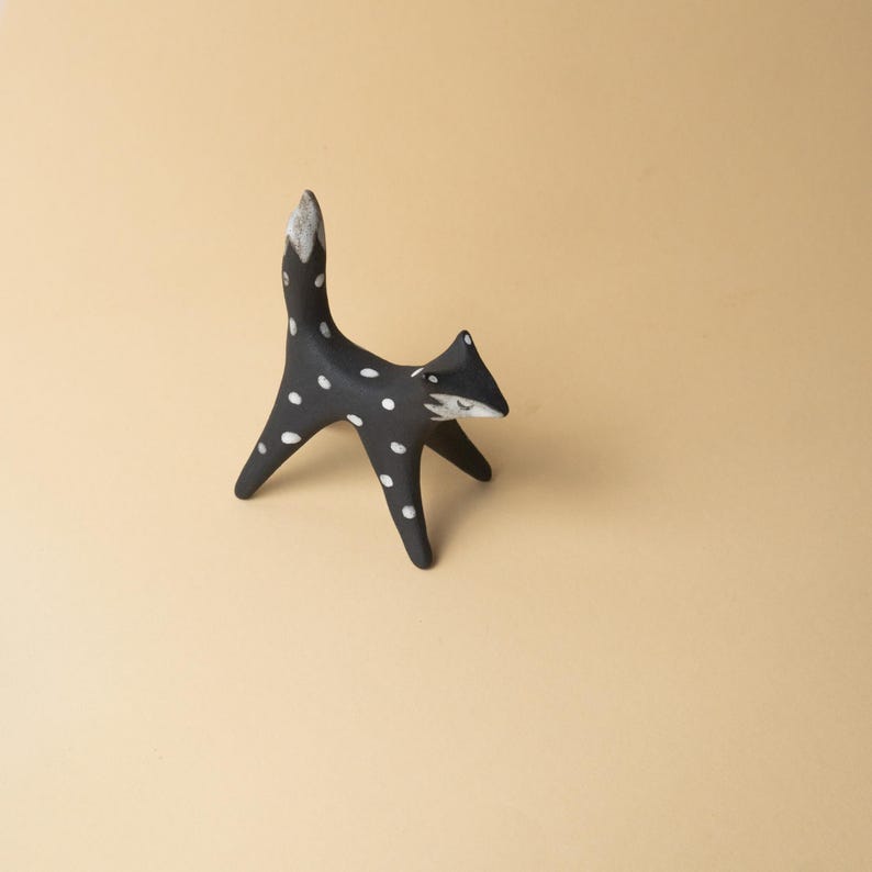 May include: A black fox ornament with white spots and a white-tipped tail. The fox has a stylized design with a pointed snout and upright posture. The ornament is set against a neutral background.